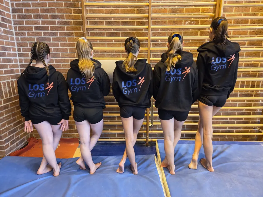 Cinq gymnastes du club, de dos, portent la veste noire LOSGYM devant les espaliers et le mur de briques de la salle Cauterman